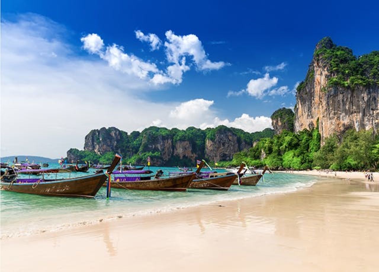 Ein Strand im Krabi Urlaub, mit hellem Sand im Vordergrund, traditionellen Booten und Felsen im Hintergrund